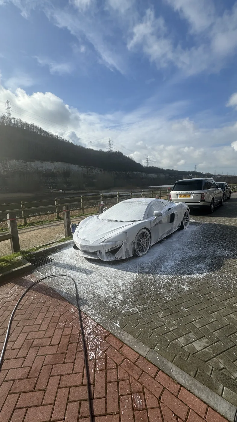 A white sports car covered in snow foam on a brick-paved drive, with a pressure washer hose in the foreground