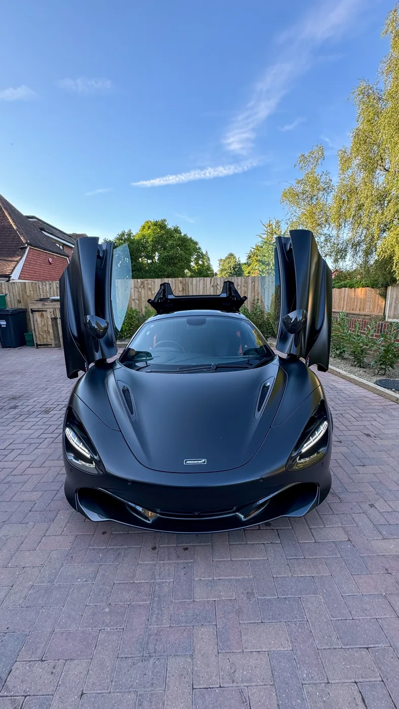 A black McLaren sports car with both doors raised, parked on a block-paved drive under a blue sky