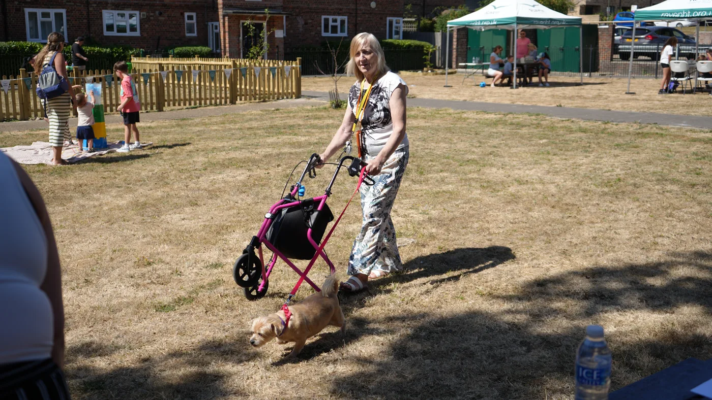 A resident with a wheeled walker leads a small dog across the grass at Nailsworth Crescent, with children playing and a Raven Housing Trust gazebo behind