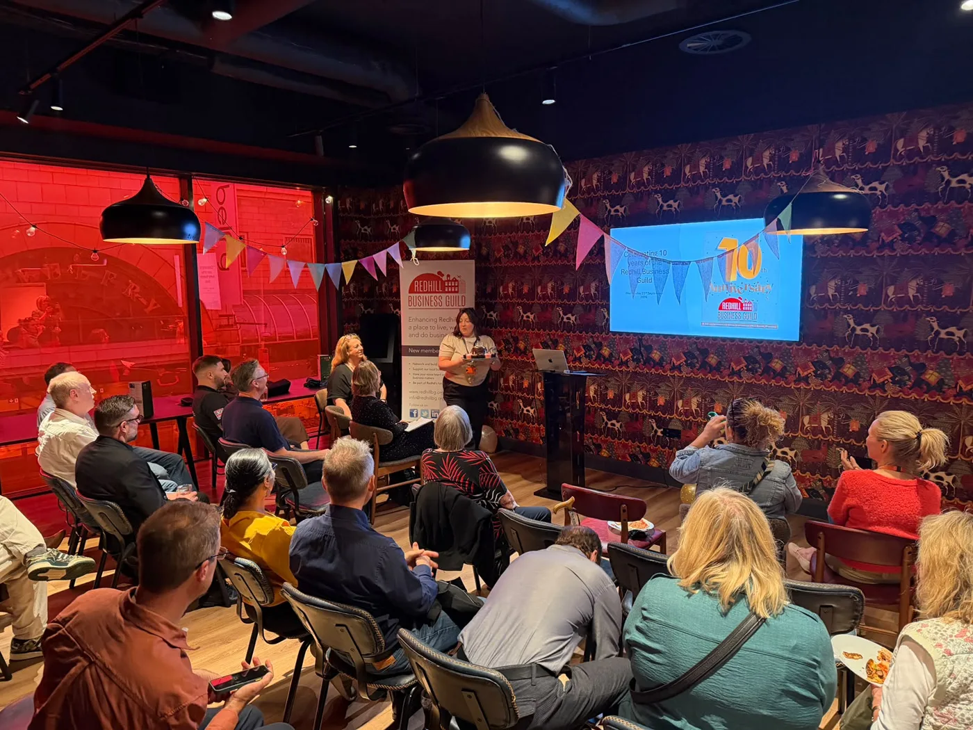 A speaker at the front of a room of seated Redhill Business Guild members, with bunting and a screen showing the number 10